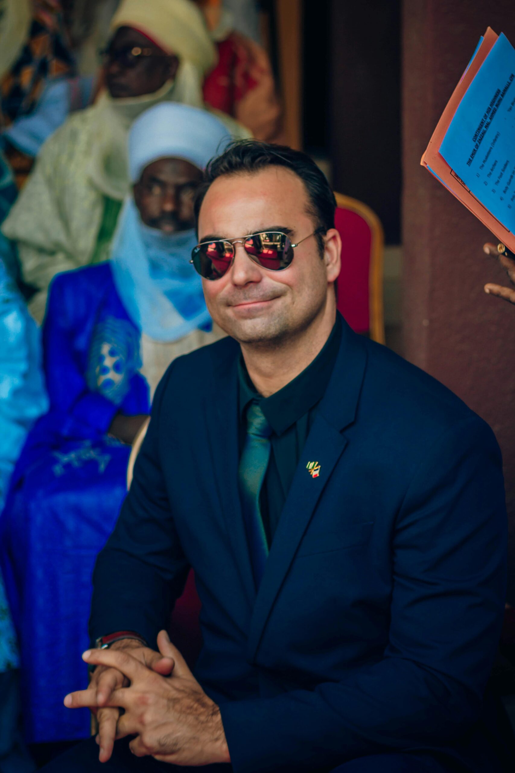Smiling man in a suit with sunglasses at a formal event with cultural attire.
